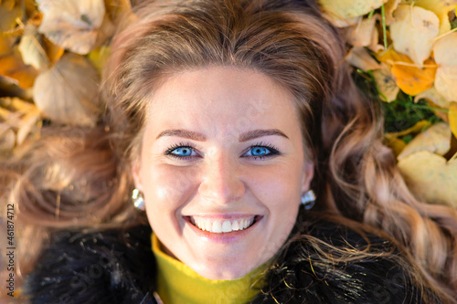 beautiful girl white toothed smile, young happy woman blue eyes whitened teeth lying in orange leaves autumn park. closeup portrait of stylish smiling lady in yellow clothes. fashion female fall 