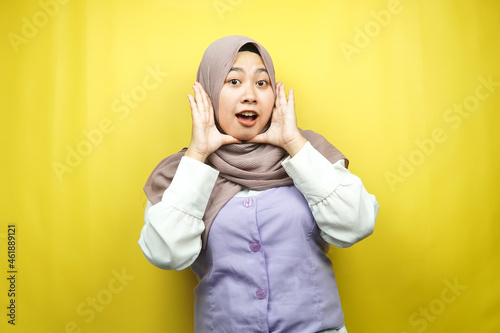 Beautiful young asian muslim woman shocked, surprised, wow expression, with hands holding cheek, isolated on yellow background © MunirSr