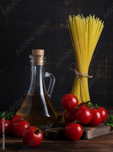 Red ripe cherry tomatoes on a green twig lie on a dark wooden board. Dry pasta spaghetti and olive oil in a jug in the background