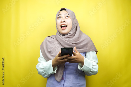 Beautiful young asian muslim woman shocked, surprised, wow expression, hand holding smartphone, looking up, isolated on yellow background