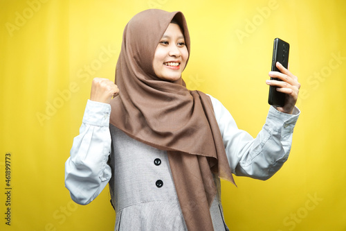 Beautiful young asian muslim woman smiling confident, enthusiastic and cheerful with hands holding smartphone, hands clenching, punching, victory, success, good work, isolated, advertising concept