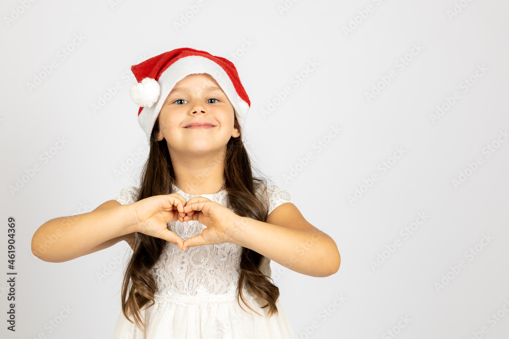 portrait of happy, gentle girl in white dress, wearing red Santa Claus hat shows heart sign from fingers isolated on white background with empty place. portrait of happy, gentle girl in white dress, wearing red Santa Claus hat shows heart sign from fingers isolated on white background with empty place.