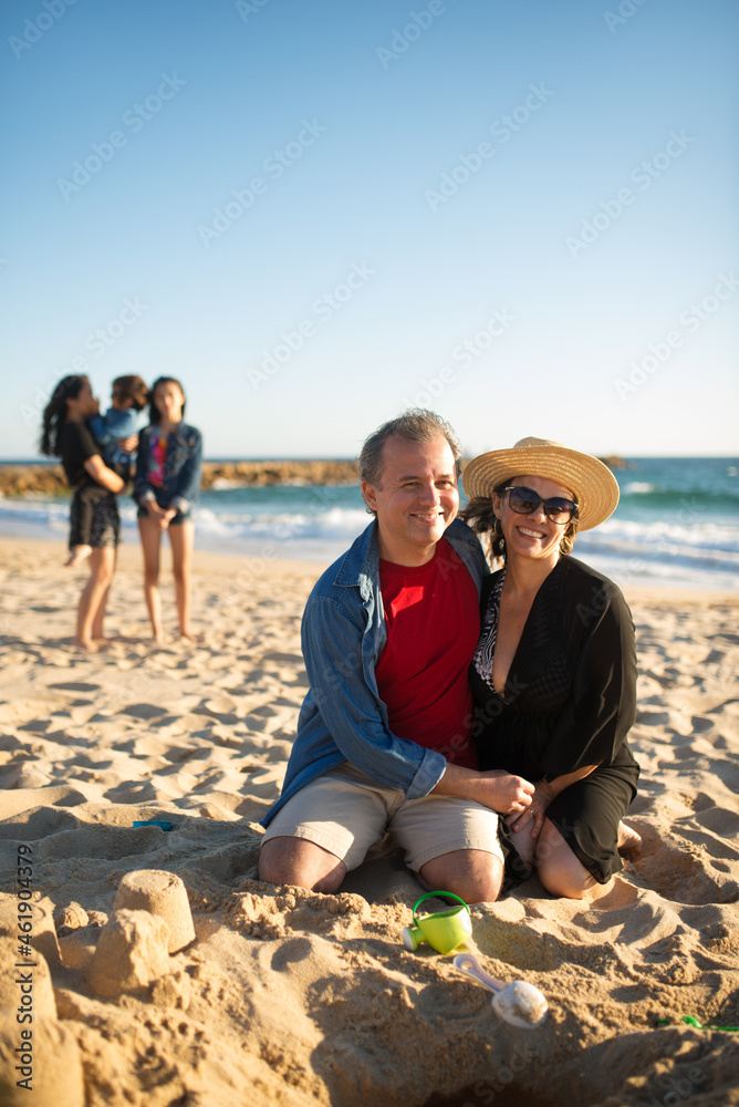 Happy smiling husband and wife at beach. Man and woman sitting at sand, hugging, looking at camera. Children in background. Family, vacation, outdoor activity concept