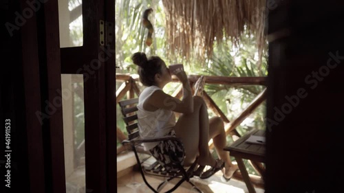 young hispanic woman drinks a coffee on the balcony in the middle of nature while holding her cell phone