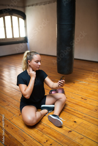 Attractive young woman checking pulse. Woman in black sport clothes sitting on floor, touching neck with fingers, counting, looking at phone. Sport, healthy lifestyle, boxing concept