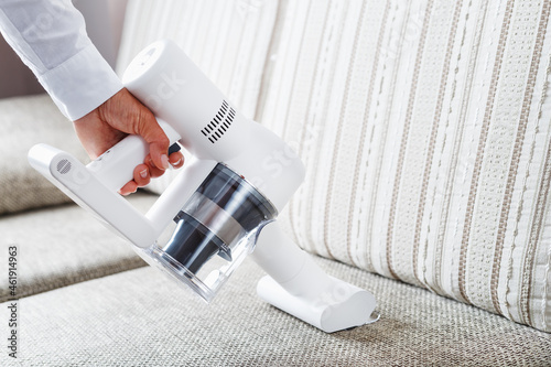 Powerful cordless vacuum cleaner with white cyclonic dust collection technology in hand, cleans the carpet in the house near the sofa.