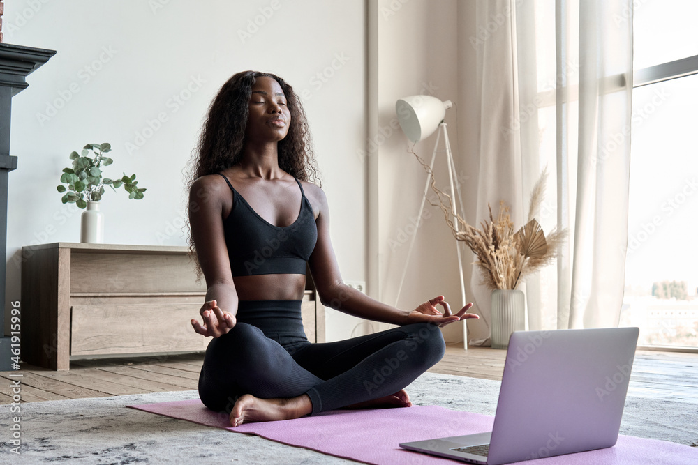 Young calm fit healthy African black woman sitting on floor at home doing  yoga breathing exercise, meditating learning online training virtual class  on computer. Exercises for mental health concept. Stock Photo