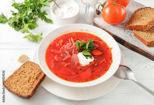red borscht with sour cream and herbs in a white plate, a spoon, bread on a white table