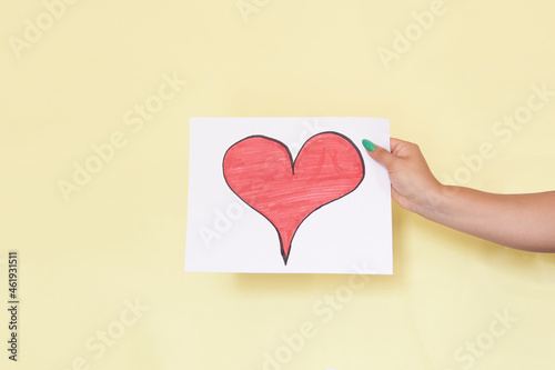 Latin woman's hand holding a sign with a red heart drawn on a white sheet, on yellow background, unrecognizable model.