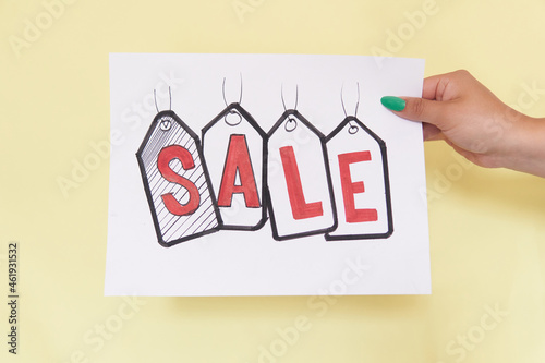 Latin woman's hand holding a sign with the word SALE written in red in drawings that look like labels on a white sheet, on yellow background, unrecognizable model.
