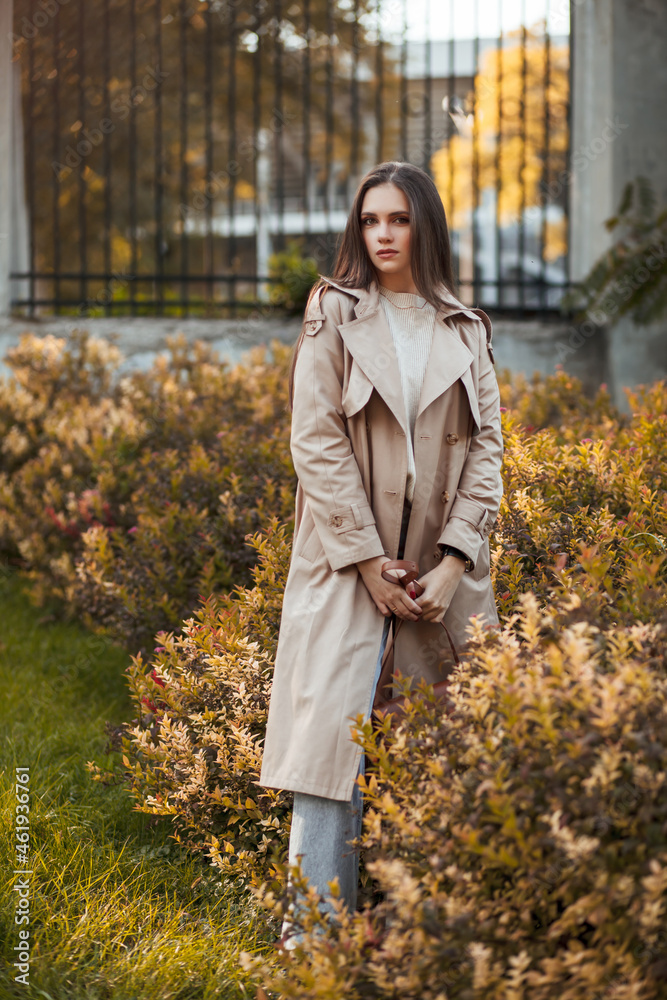 A beautiful tall girl of model appearance in a beige raincoat and gray jeans walks in a city park on a warm autumn day.Lifestyle concept. Autumn street fashion look. A beautiful tall girl of model appearance in a beige raincoat and gray jeans walks in a city park on a warm autumn day.Lifestyle concept. Autumn street fashion look.