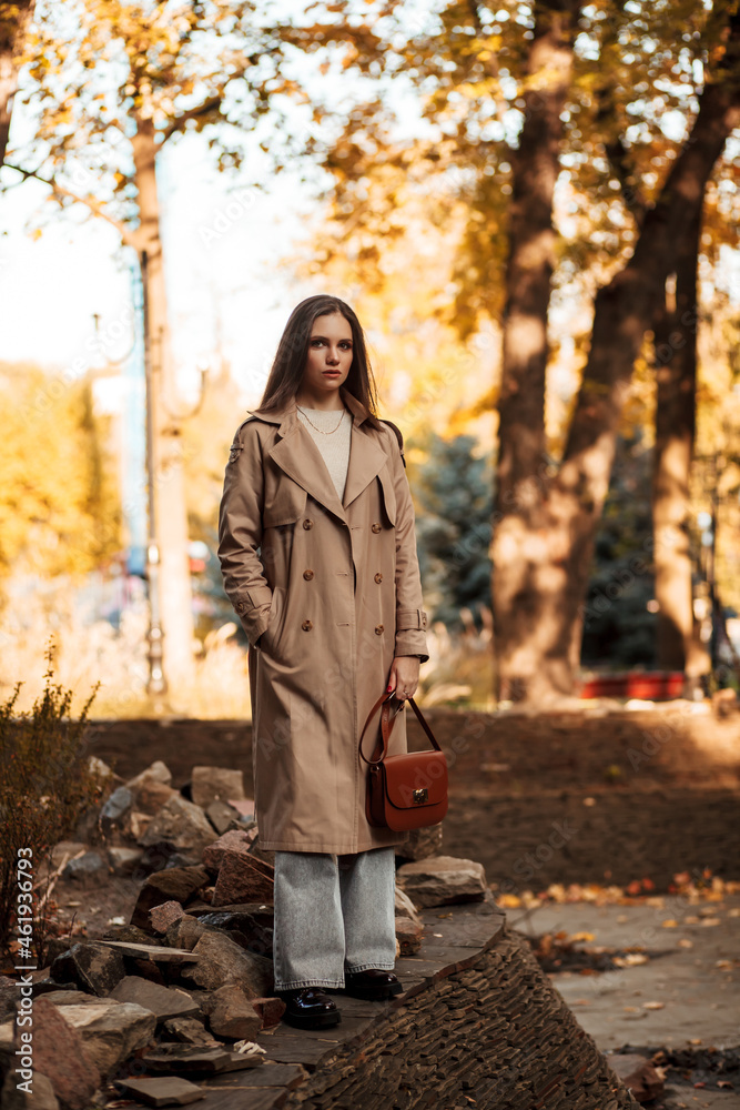 A beautiful tall girl of model appearance in a beige raincoat and gray jeans walks in a city park on a warm autumn day.Lifestyle concept. Autumn street fashion look. A beautiful tall girl of model appearance in a beige raincoat and gray jeans walks in a city park on a warm autumn day.Lifestyle concept. Autumn street fashion look.