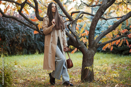 A beautiful tall girl of model appearance in a beige raincoat and gray jeans walks in a city park on a warm autumn day.Lifestyle concept. Autumn street fashion look.