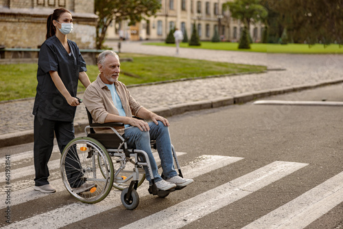 Full length shot of young nurse in protective face mask taking care of mature disabled man in wheelchair during a walk in the city