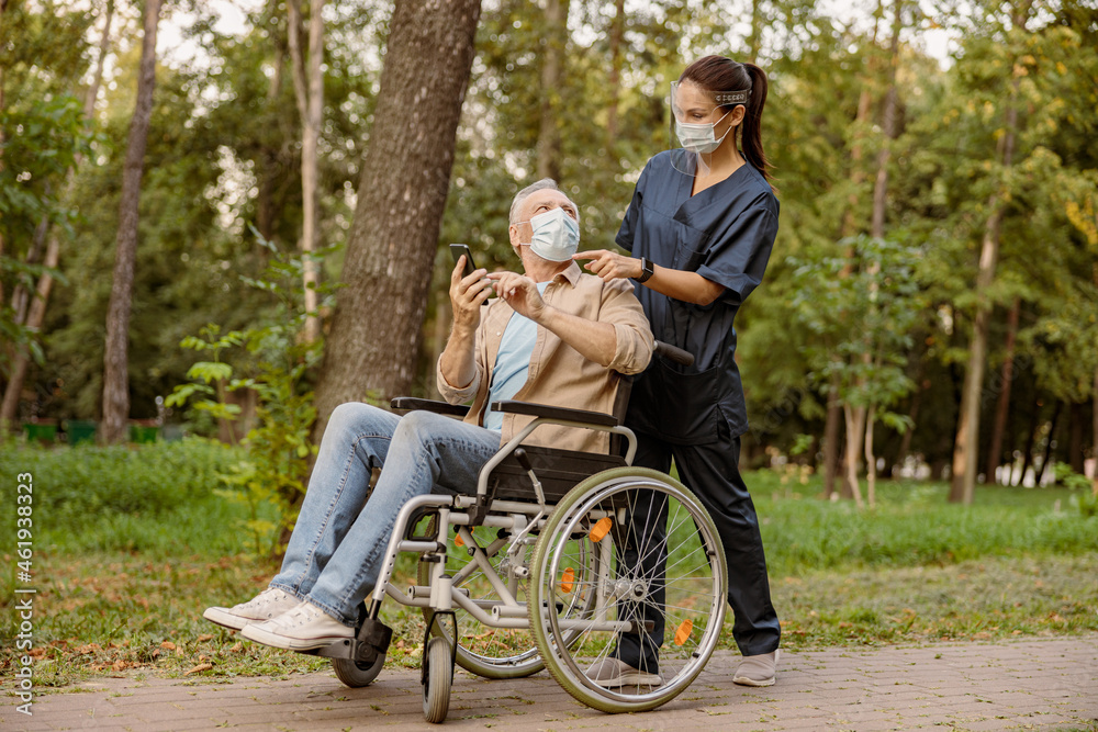 Young nurse in face shield and protective face mask assisting senior handicapped man in wheelchair using his smartphone during a walk in the park Young nurse in face shield and protective face mask assisting senior handicapped man in wheelchair using his smartphone during a walk in the park