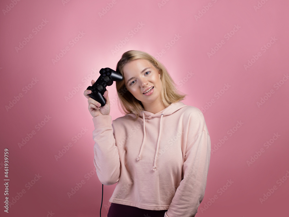A gamer who is passionate about the process. A cute blonde is holding a gamepad in her hands, looking at the camera. A young woman in a pink hoodie, holding a joystick in her hands ,photo on pink A gamer who is passionate about the process. A cute blonde is holding a gamepad in her hands, looking at the camera. A young woman in a pink hoodie, holding a joystick in her hands ,photo on pink