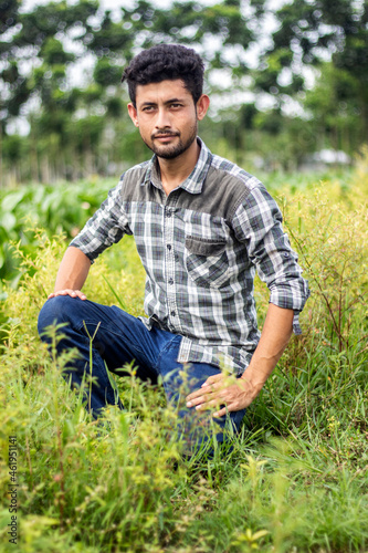 A handsome boy is sitting in the middle of the green nature in a grey shirt and has a small beard on his face. the background blur. © Rokonuzzamnan