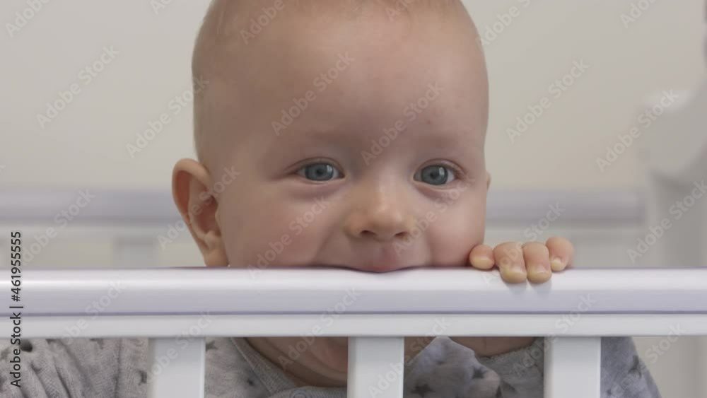 Baby boy sitting in white wooden crib holding hands on the rail of rocking cradle, face of cute adorable infant closeup. High quality 4k footage Baby boy sitting in white wooden crib holding hands on the rail of rocking cradle, face of cute adorable infant closeup. High quality 4k footage