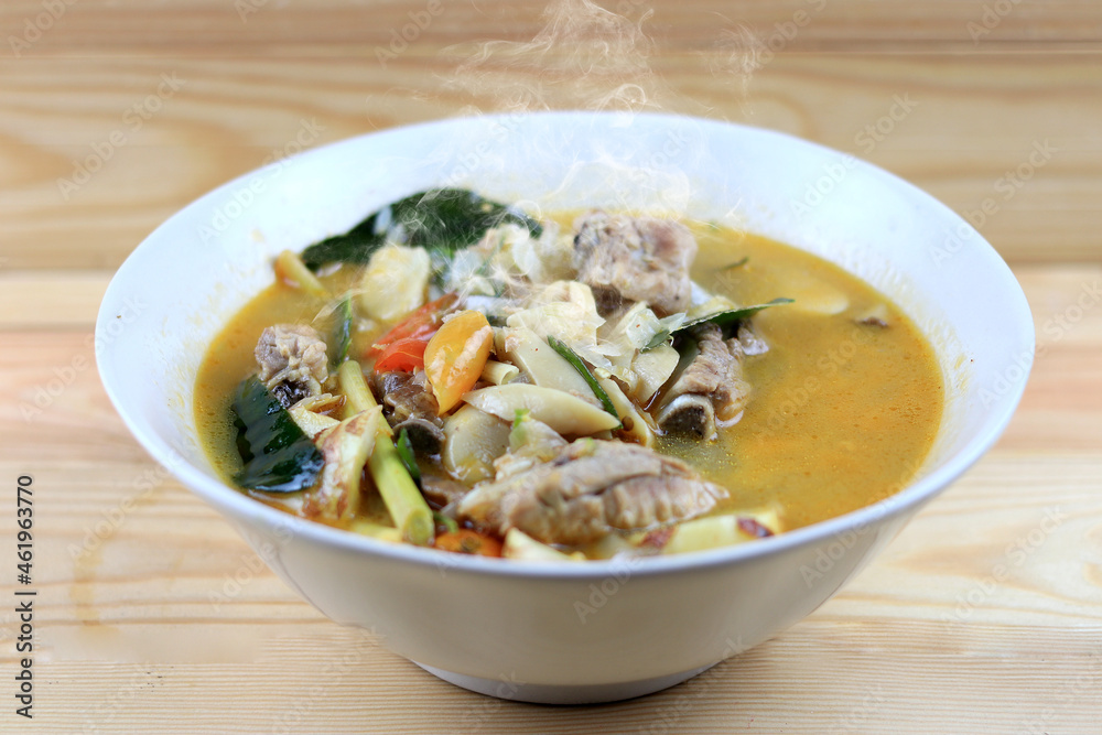 Spicy boiled Pork bone and straw mushroom, wild tomato, herb in tom yum soup on white bowl on wooden table. Tom Yum soup pork in Thai style food. Spicy boiled Pork bone and straw mushroom, wild tomato, herb in tom yum soup on white bowl on wooden table. Tom Yum soup pork in Thai style food.