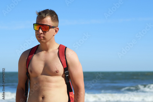 Young athletic guy looks to the side in sunglasses on the beach. Vacation and recreation on the seaside. Sunny bright day on a journey by the sea.