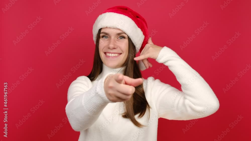 Happy young woman in white cozy sweater and Santa Christmas hat doing phone gesture like says call me back, posing isolated on red background studio. Happy New Year celebration merry holiday concept