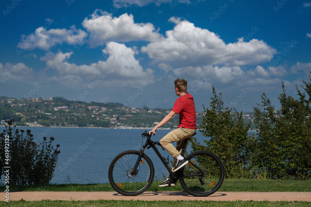 Obraz premium The movement of a cyclist in a red T-shirt along the coastline in the background lake, mountains, blue sky with clouds. A man on a mountain bike lake garda.