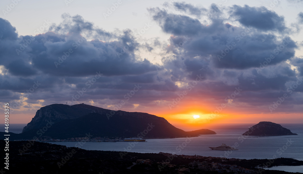 Stunning panoramic view of a coastline with the island of Figarolo and a luxury yacht sailing on a calm water. Beautiful and dramatic sunrise during a cloudy day, Golfo Aranci, Sardinia, Italy.