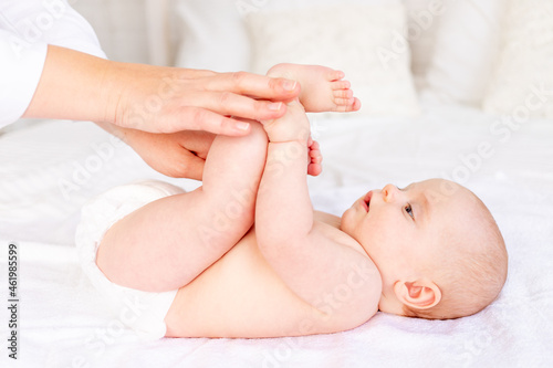 mom's hands hold the baby by the legs while doing massage or gymnastics in the crib on a white cotton bed, hygiene and care of the newborn