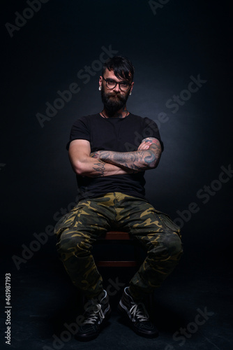 Brutal attractive bearded man with tattooed hands poses in black blank t-shirt and army pants seated on a chair © qunica.com
