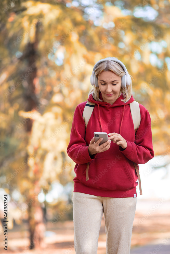 Digital zombie. Adult woman in big headset go through the park and stick to her mobile, outdoor in autumn, vertical photo, selected focus. Digital zombie. Adult woman in big headset go through the park and stick to her mobile, outdoor in autumn, vertical photo, selected focus.