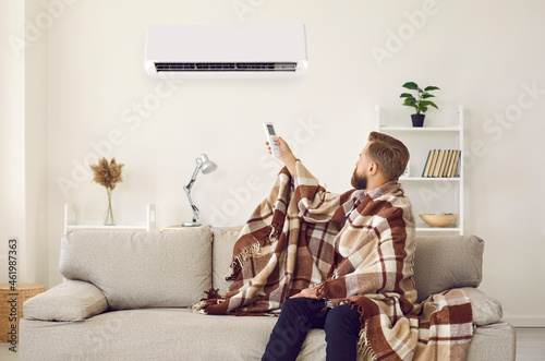 Sad angry man sitting on sofa trying to warm up under plaid while freezing in cold living room with modern air conditioner turned on. Young guy having trouble adjusting AC system temperature at home