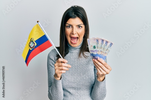 Young hispanic woman holding colombia flag and colombian pesos banknotes celebrating crazy and amazed for success with open eyes screaming excited.