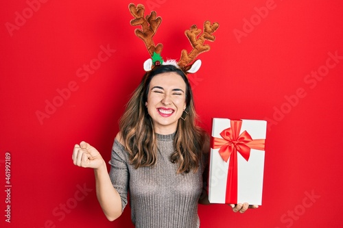 Young hispanic girl wearing deer christmas hat holding gift screaming proud, celebrating victory and success very excited with raised arm