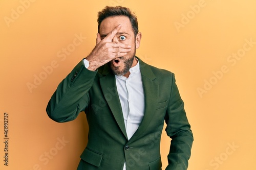 Middle age man wearing business suit peeking in shock covering face and eyes with hand, looking through fingers with embarrassed expression.