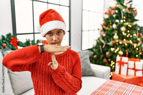 Young hispanic woman with short hair wearing christmas hat sitting on the sofa doing time out gesture with hands, frustrated and serious face