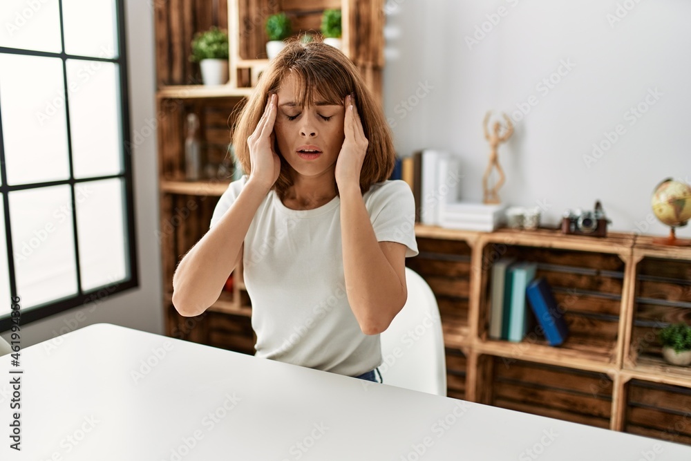 Young caucasian girl wearing casual clothes sitting on the table at home with hand on head for pain in head because stress. suffering migraine.