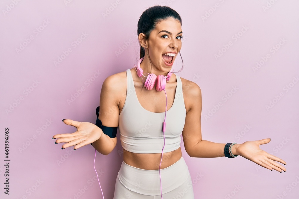 Young hispanic woman wearing gym clothes and using headphones smiling cheerful with open arms as friendly welcome, positive and confident greetings