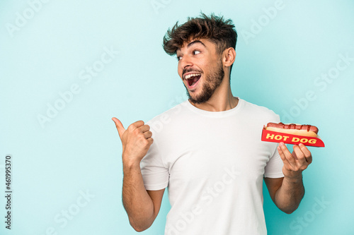 фотографія Young arab man holding a hotdog isolated on blue background points with thumb finger away, laughing and carefree