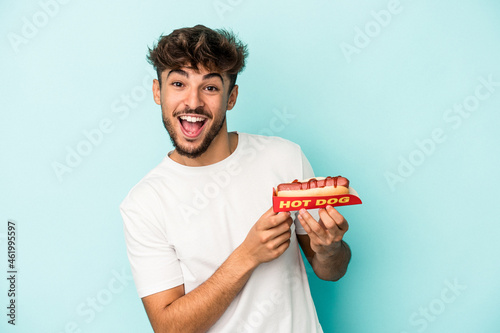 Young arab man holding a hotdog isolated on blue background laughing and having fun.