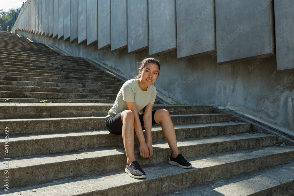 Smiling Asian sportswoman in tracksuit sits on underground crossing stone stairs by wall resting during training in morning