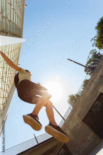 Strong sportive woman in tracksuit with sneakers jumps high up training on city street under clear blue sky close bottom view