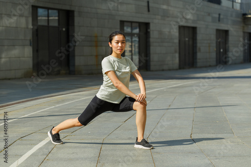 Positive strong Asian woman in tracksuit and shoes does forward dynamic lunges training on sunny city square on summer day