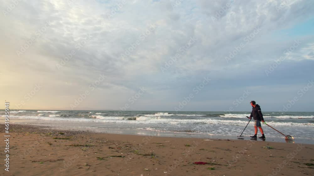 A man is looking for the lost luxuries with electronic metal detector and dragging a scoop behind him on the beach. Nice fresh morning and blue sky above the sea A man is looking for the lost luxuries with electronic metal detector and dragging a scoop behind him on the beach. Nice fresh morning and blue sky above the sea