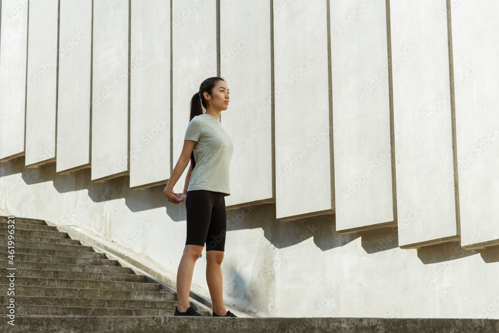 Obraz premium Pretty young Asian woman in stylish tracksuit stretches arms behind back near wall with concrete decor on sunny city street