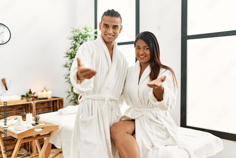 Young latin couple wearing towel standing at beauty center smiling friendly offering handshake as greeting and welcoming. successful business.