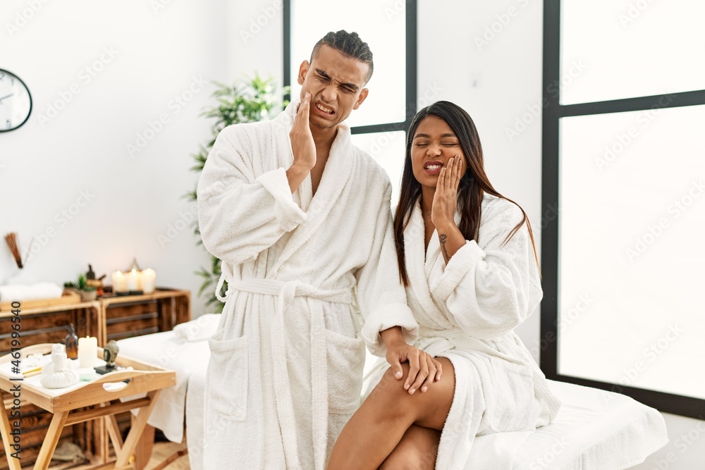 Young latin couple wearing towel standing at beauty center touching mouth with hand with painful expression because of toothache or dental illness on teeth. dentist concept.