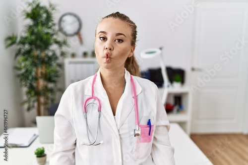 Young caucasian woman wearing doctor uniform and stethoscope at the clinic making fish face with lips, crazy and comical gesture. funny expression.