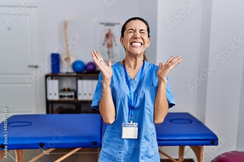 Young hispanic woman wearing physiotherapist uniform standing at clinic celebrating mad and crazy for success with arms raised and closed eyes screaming excited. winner concept