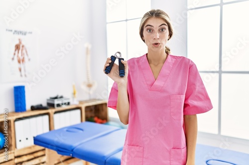 Young blonde woman working at pain recovery clinic holding hand strengthener scared and amazed with open mouth for surprise  disbelief face