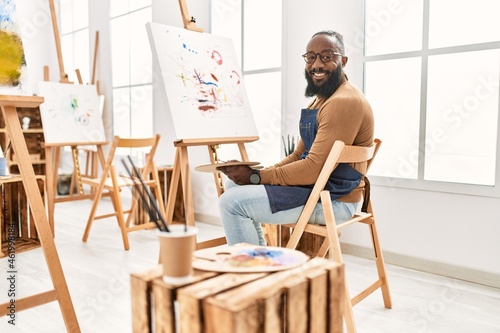African american artist man painting on canvas at art studio looking positive and happy standing and smiling with a confident smile showing teeth
